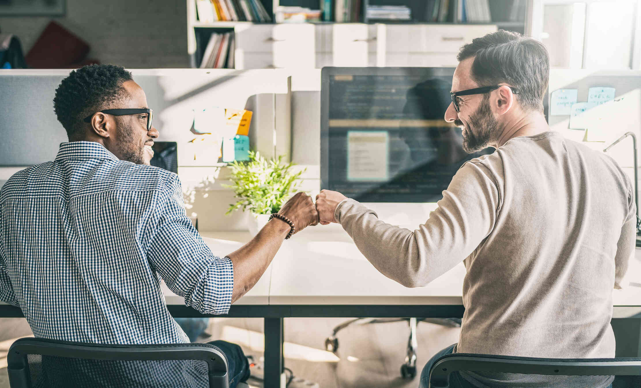 Two men wearing glasses sit next to each other at desks in front of desktop computers. They look at each other, smile, and fist bump.